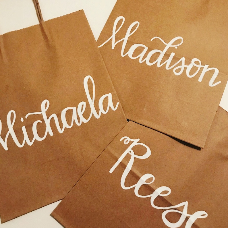 May include: Four brown paper gift bags with white cursive lettering. The names "Michaela", "Madison", and "Reese" are visible. The bags have a natural, rustic appearance, with a twisted paper handle on one bag.