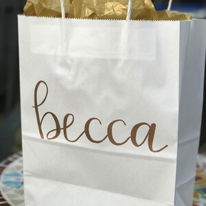 May include: White paper gift bag with the name "becca" in a brown script font. The bag contains gold tissue paper and has white rope handles.