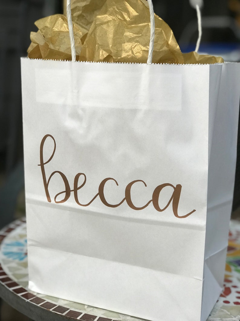 May include: White paper gift bag with the name "becca" in a brown script font. The bag contains gold tissue paper and has white rope handles.