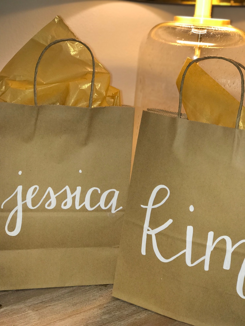 May include: Two brown paper gift bags with white cursive lettering that reads "jessica" and "kim". Each bag has a twisted paper handle and is filled with gold tissue paper. The background includes a glass lamp.