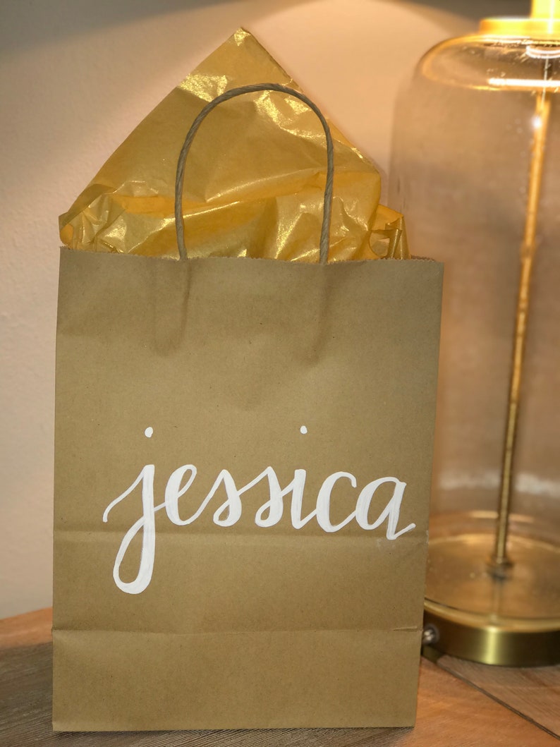 May include: A brown paper gift bag with the name "jessica" in white script. The bag contains gold tissue paper and has a twisted paper handle. The bag is sitting on a wooden surface.