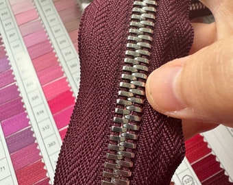 Op de afbeelding: Close-up van een bordeauxrode rits met een zilveren metalen rits. De rits ligt op een wit oppervlak met kleurstalen.