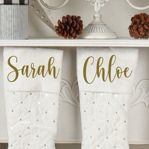 May include: Three white Christmas stockings with gold glitter and names "Sarah", "Chloe", and "Noah" written on them.