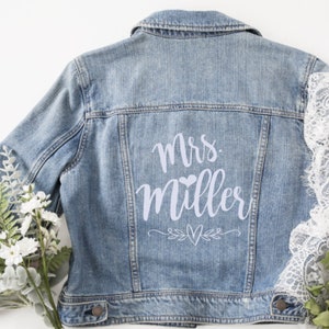 May include: A light blue denim jacket with the text "Mrs. Miller" in white lettering on the back. The lettering is in a cursive font with a small heart above the last name. There are two small hearts and a floral design below the name.