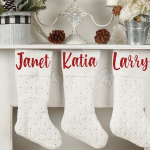 May include: Three white Christmas stockings with red glitter names, "Janet", "Katia", and "Larry", hanging on a white mantel.