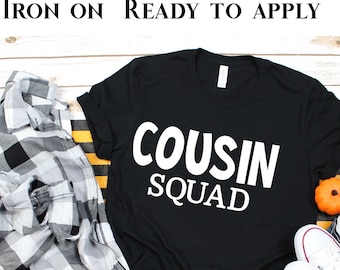 May include: Black t-shirt with white text that reads "COUSIN SQUAD". The shirt is laid out on a white wooden surface with a plaid shirt, jeans, and a pair of white sneakers.