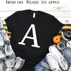 May include: Black t-shirt with a white letter A iron-on design. The shirt is laid flat on a wooden surface with a plaid shirt, jeans, and Halloween decorations.