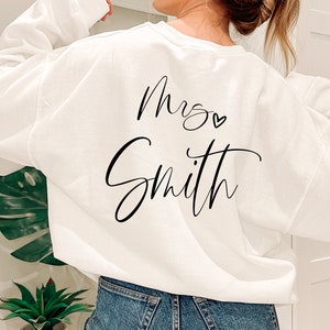 May include: White sweatshirt with the text "Mrs. Smith" in black script. A small heart is above the name. The sweatshirt is a casual style, perfect for a wedding or bridal event.