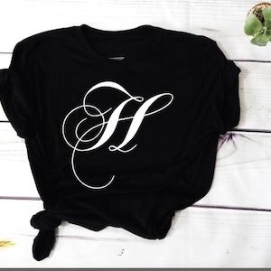 May include: Black t-shirt with a white cursive letter H on the front.