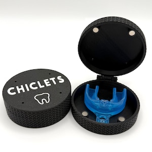 May include: A black circular case with a textured exterior and the word CHICLETS printed in white above a tooth icon. The case is open, revealing a translucent blue dental guard inside, with small circular magnets visible on the inner rim.