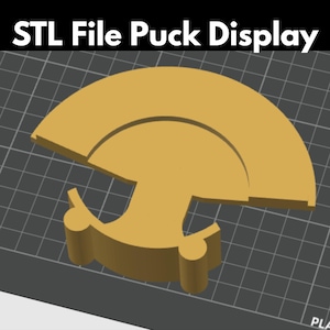 May include: A digital 3D model of a tan puck display stand featuring a semi-circular top section and a base with two cylindrical supports. The text "STL File Puck Display" is visible at the top of the image against a dark gray grid background.
