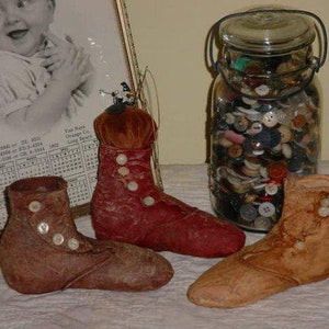 May include: Three vintage fabric boots with button accents. The boots are brown, red, and tan. A glass jar filled with buttons is in the background.