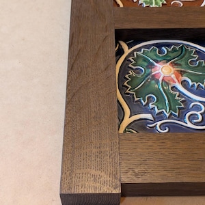 May include: Close-up of a decorative wooden frame with colorful ceramic tiles. The tiles feature a floral design with green leaves, red accents, and a blue background. The frame is made of dark brown wood.
