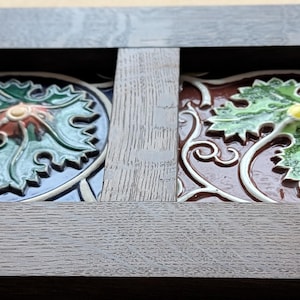 May include: Close-up of two decorative tiles with floral designs. One tile has a blue background with a green and red floral pattern. The other tile has a brown background with a green and yellow floral pattern. Both tiles are framed in dark wood.