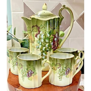 May include: A vintage tea set featuring a large teapot and four matching mugs. The set is painted with a green and purple grape design. The teapot has a tall, curved handle and a lid with a small finial. The mugs have a scalloped edge.