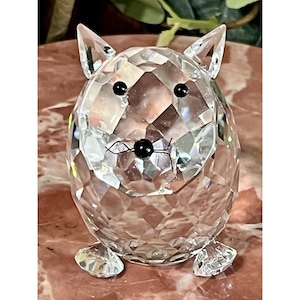 May include: A clear, faceted crystal figurine of a cat. The cat has black eyes and a black nose, with whiskers. The figurine is sitting on a pink surface. The cat has pointed ears and small feet.
