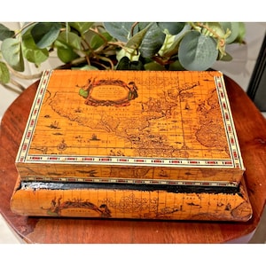 May include: A rectangular, antique-style wooden box with a hinged lid. The box is decorated with a detailed map design in shades of brown and tan, with decorative borders. The box is sitting on a wooden surface.