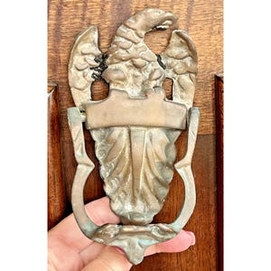 May include: Antique brass eagle door knocker. The door knocker is mounted on a wooden door. The eagle has outstretched wings and a banner across the chest. The knocker is held in place by a curved handle.