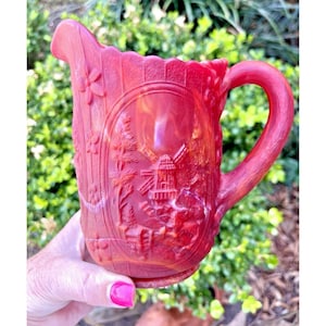 May include: A red glass pitcher with a handle and spout. The pitcher features a detailed embossed design of a windmill, trees, and flowers. The pitcher has a scalloped rim and is held by a person.