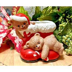 May include: A ceramic Christmas decoration featuring two teddy bears. One bear wears a Santa hat and peeks over the top of a red boot. The other bear rests on the boot. The boot has a white fluffy trim and a black base.