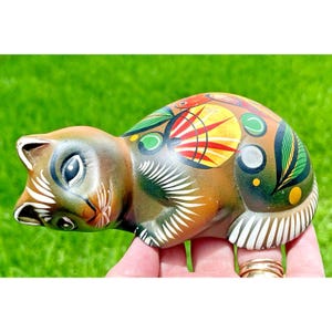 May include: A hand-painted ceramic cat figurine in shades of brown, green, yellow, and red. The cat is lying down with detailed patterns on its back and sides. The figurine has white accents on its face and paws.