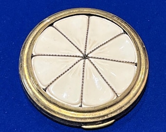 May include: A round, vintage compact with a gold-toned metal frame. The top features a white enamel design divided into eight segments by thin, dark lines. The compact is set against a blue background.