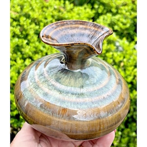 May include: A handcrafted ceramic vase with a unique, organic shape. The vase features a round body and a flared, petal-like opening. The glaze has a blend of brown, green, and blue hues, creating a marbled effect. The vase is held in a hand.