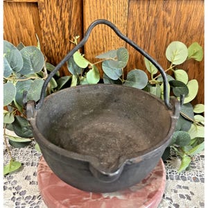 May include: A small, dark gray cast iron cauldron with a curved handle. The cauldron has a spout and is sitting on a pink and white marble-patterned surface. The background is a wooden surface with green leaves.