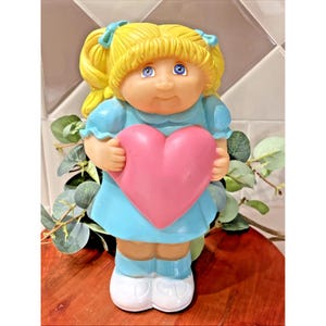 May include: A vintage Cabbage Patch Kids doll figurine. The doll has blonde pigtails, blue eyes, and is wearing a light blue dress. It is holding a pink heart. The doll is standing on a wooden surface.