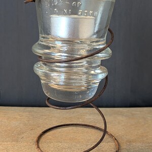 May include: A clear glass insulator with a wire coil around it, sitting on a rusty metal spring base. The insulator has the text "MADE IN U.S.A. 2-98 - 60" printed on it.