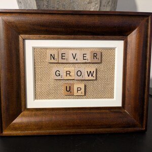 May include: A framed piece of art with the words "NEVER GROW UP" spelled out using letter tiles on a burlap background.