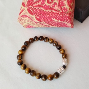 Könnte beinhalten: Ein braunes Tigerauge-Armband mit zwei silbernen Buddha-Kopf-Anhängern.