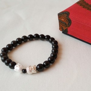 Könnte beinhalten: Ein schwarzes Perlenarmband mit zwei silbernen Buddha-Kopf-Anhängern. Das Armband liegt auf einer weißen Oberfläche neben einer roten Schachtel mit Blumenmuster.