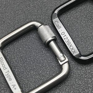 May include: Two square carabiner clips, one silver and one black, with screw-lock closures. The silver clip is engraved with "Blood Type: A+", and the black clip has "Call: 01239XXX". Both are on a textured grey surface.