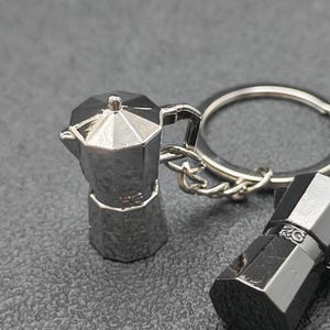 Op de afbeelding: Een zilverkleurige metalen sleutelhanger met een miniatuur koffiezetapparaat. De sleutelhanger heeft een gedetailleerd ontwerp, inclusief een handvat en een tuit. Het koffiezetapparaat is bevestigd aan een zilveren sleutelring. De achtergrond is een getextureerd grijs oppervlak.
