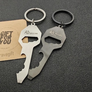 Pode incluir: Dois chaveiros com abridor de garrafas. Um é prateado, o outro cinza escuro. Ambos têm um design em forma de chave com as iniciais "AD" e uma data gravada. Um pequeno cartão com o texto "Gift for You" também é visível.