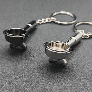Könnte beinhalten: Zwei silberfarbene Schlüsselanhänger mit Siebträger für Espressomaschinen. Jeder Schlüsselanhänger hat einen Miniatur-Siebträger mit detailliertem Design, der an einer Kette und einem Schlüsselring befestigt ist. Die Schlüsselanhänger werden auf einer dunklen Oberfläche präsentiert.