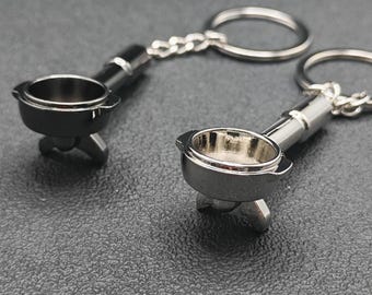 以下が含まれることがあります： 2つのシルバーカラーのエスプレッソマシン用ポルタフィルターキーホルダー。各キーホルダーは、チェーンとキーリングに取り付けられた、詳細なデザインのミニチュアポルタフィルターを備えています。キーホルダーは暗い表面に表示されています。