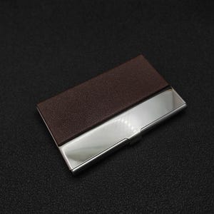 A brown leather wallet and a silver metal card holder are placed on a dark surface.