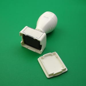 Può includere: Un timbro autoinchiostrante bianco con un vano rettangolare per il tampone e un coperchio staccato, su sfondo verde. L'impugnatura del timbro è arrotondata e il testo "FS 05" è visibile in alto.