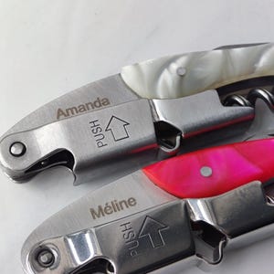 Może przedstawiać: Dwa korkociągi kelnerskie ze stali nierdzewnej ze składanymi mechanizmami. Jeden uchwyt ma białą masę perłową i imię Amanda, a drugi jaskraworóżową masę perłową i imię Méline. Oba posiadają strzałkę z napisem PUSH.
