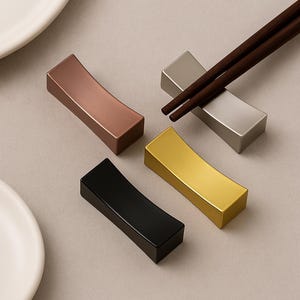 May include: Four metallic chopstick rests in copper, silver, gold, and black. One silver rest holds a pair of brown chopsticks. The rests have a curved, rectangular shape and are arranged on a neutral gray surface.