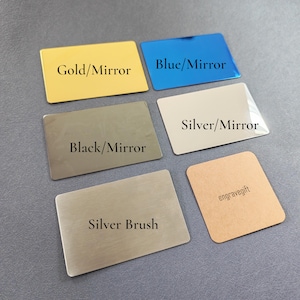 Può includere: Un assortimento di carte metalliche rettangolari in vari colori e finiture. Le carte includono finiture a specchio oro, blu, nero e argento e una finitura spazzolata argento. Una carta è marrone chiaro con il testo "engravegift".