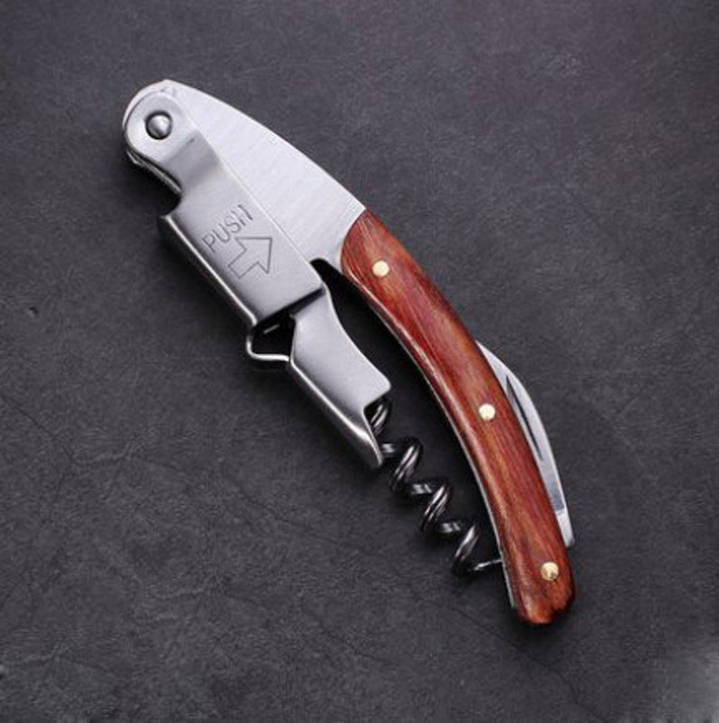 May include: A silver and wood corkscrew with a  "PUSH" label and an arrow pointing upwards. The corkscrew has a spring-loaded worm and a lever.