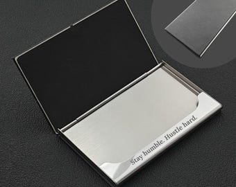 May include: A silver business card holder with a brushed metal finish, open to reveal a black interior. The phrase "Stay humble. Hustle hard." is engraved on the front. A smaller, closed holder is visible in the background.