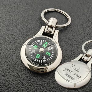 Op de afbeelding: Zilveren kompas sleutelhanger met een zwart en groen kompas gezicht. De sleutelhanger heeft een ring voor sleutels en een decoratieve achterkant met de woorden "Find your way home".
