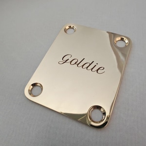 May include: A polished gold guitar neck plate with four mounting holes. The word "Goldie" is elegantly engraved in a cursive font across the plate's surface. The plate has a rectangular shape with rounded corners.
