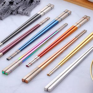 Może przedstawiać: Zestaw ośmiu par pałeczek w różnych kolorach, w tym czarny, czerwony, niebieski, tęczowy, pomarańczowy, złoty i srebrny. Pałeczki są wykonane z metalu i mają polerowane wykończenie.