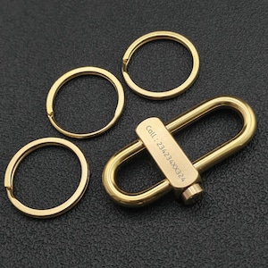 Puede incluir: Un llavero dorado con una barra rectangular y un cierre en forma de U, acompañado de tres llaveros. La barra está grabada con el texto "Call: 234234XX324". Los artículos son de metal y están sobre una superficie oscura.