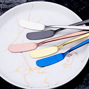 Puede incluir: Cinco cuchillos para mantequilla en un plato blanco con un patrón de mármol. Los cuchillos están hechos de acero inoxidable y tienen mangos de diferentes colores: plateado, negro, oro rosa, oro y azul.